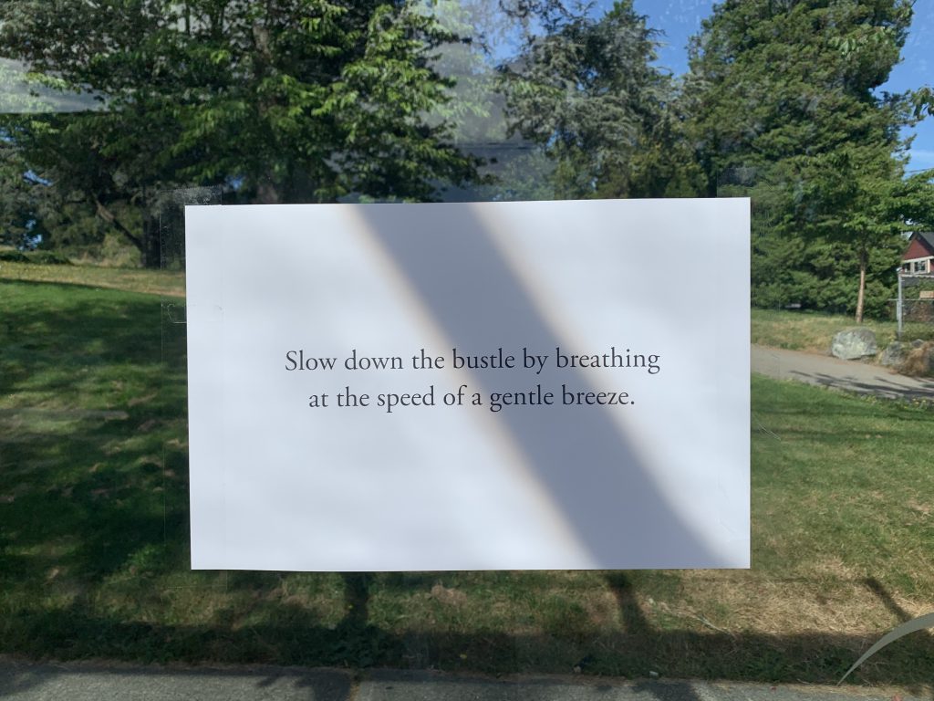 A piece of paper on a glass surface with nature in the background. The paper has the following words on it: Slow down the bustle by breathing at the speed of a gentle breeze. Photo by Alyssa Martens